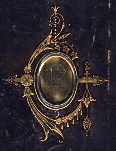 Emblema familia Pérez-Villamil, en forma de óvalo de metal dorado con las iniciales PV grabadas, rodeado por una orla modernista dorada