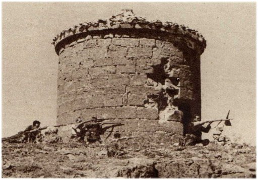 Imagen en blanco y negro del molino en agosto de 1936, con cuatro milicianos agazapados haciendo ademán de disparar en dirección sur. A la derecha, aparece un banderín, sujeto sobre lo que aparentan ser un montón de piedras a modo de mojón