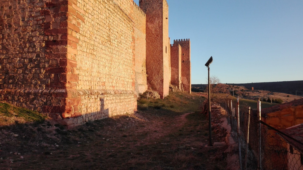 Camino flanqueado por un muro bajo a la izquierda y las murallas del castillo a la derecha, terminando en el muro trasero del Hostal de Doña Blanca