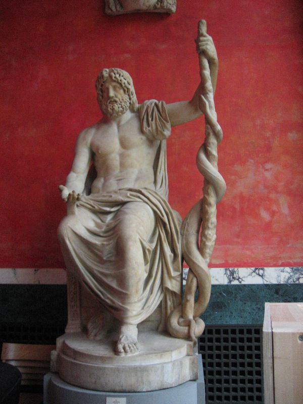 Una representación del héroe divinizado Asklepios, sentado, con el torso desnudo, en la que porta una vara en su mano izquierda, en torno a la cual se enrosca una serpiente de grandes dimensiones